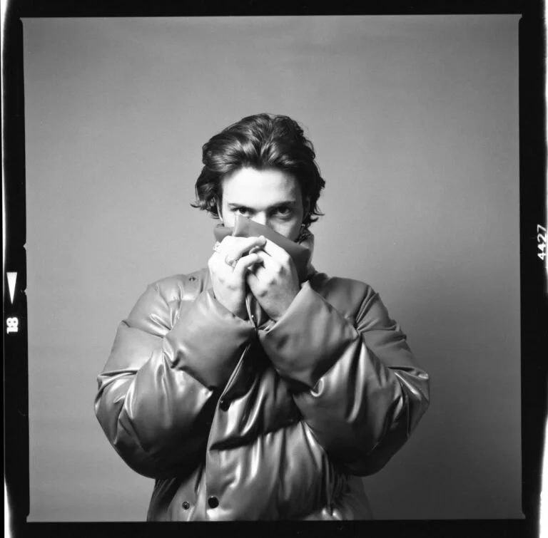 Jeune homme en manteau, tenant une écharpe devant la bouche, regardant intensément en camera, photo en noir et blanc.