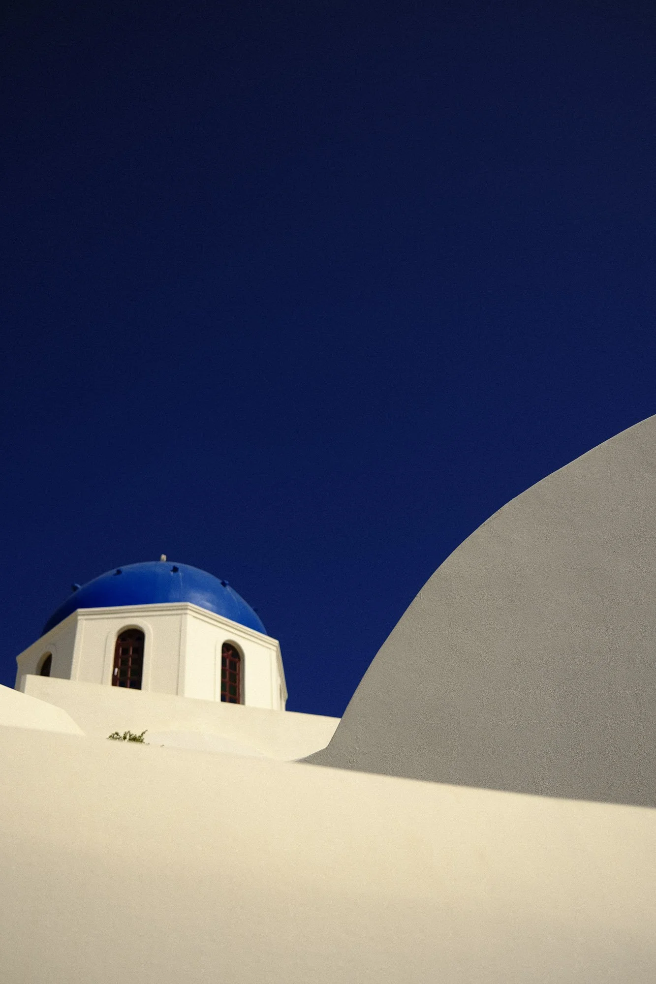 Une église blanche avec un dôme bleu, typique de l'architecture grecque, sous un ciel bleu profond. La photo est prise d'un angle incluant des éléments d'architecture blanche au premier plan.