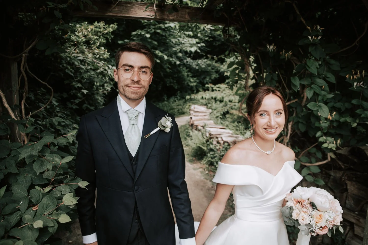 Un couple de mariés en costume et robe blanche, tenant une bouquet de fleurs, posent dans un jardin verdoyant lors de leur mariage.