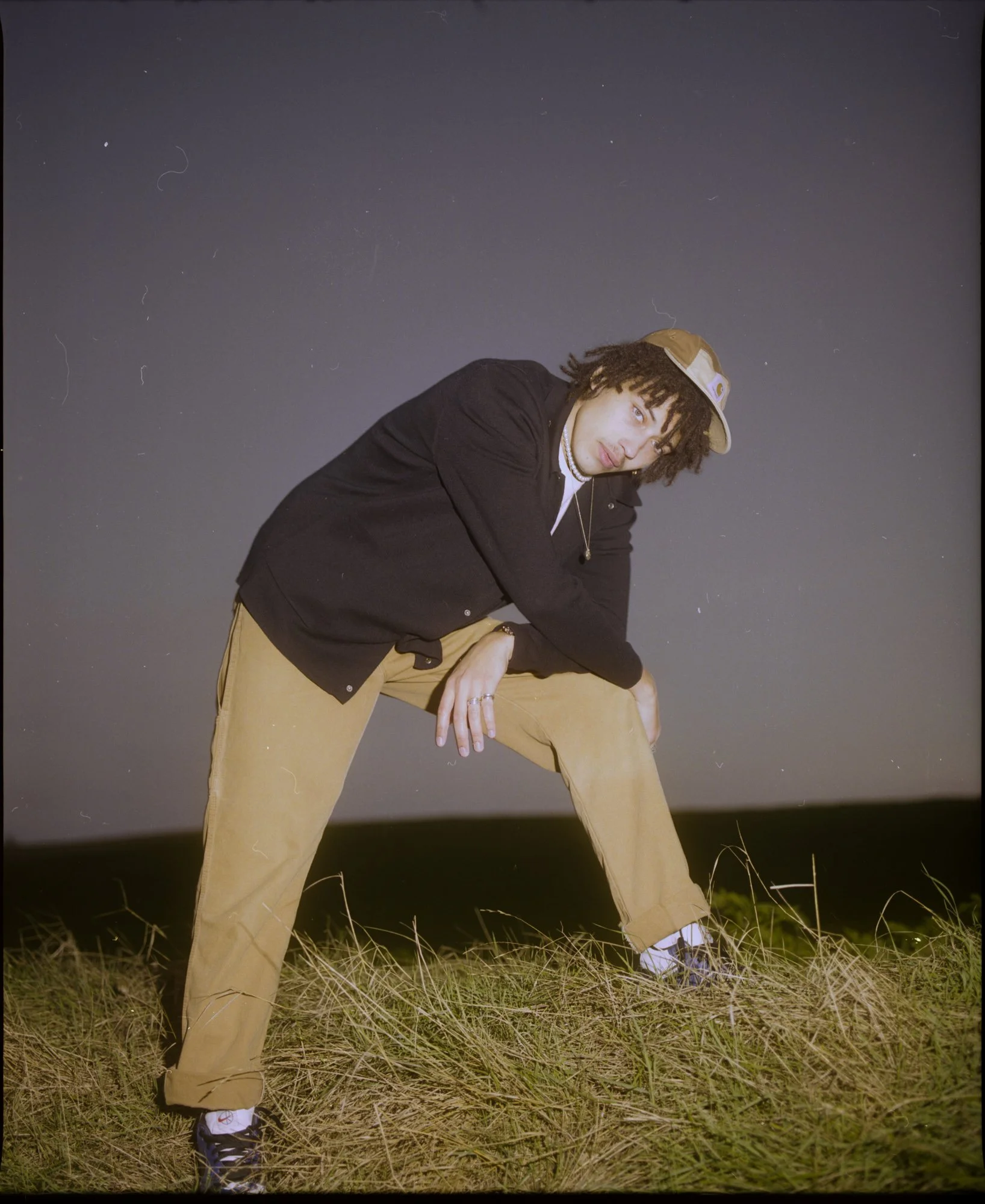 Jeune homme debout dans un champ avec herbes hautes, portant un chapeau de baseball beige, un sweat noir, un collier de perles et des pantalons beige, vu de côté avec un ciel nocturne en arrière-plan.