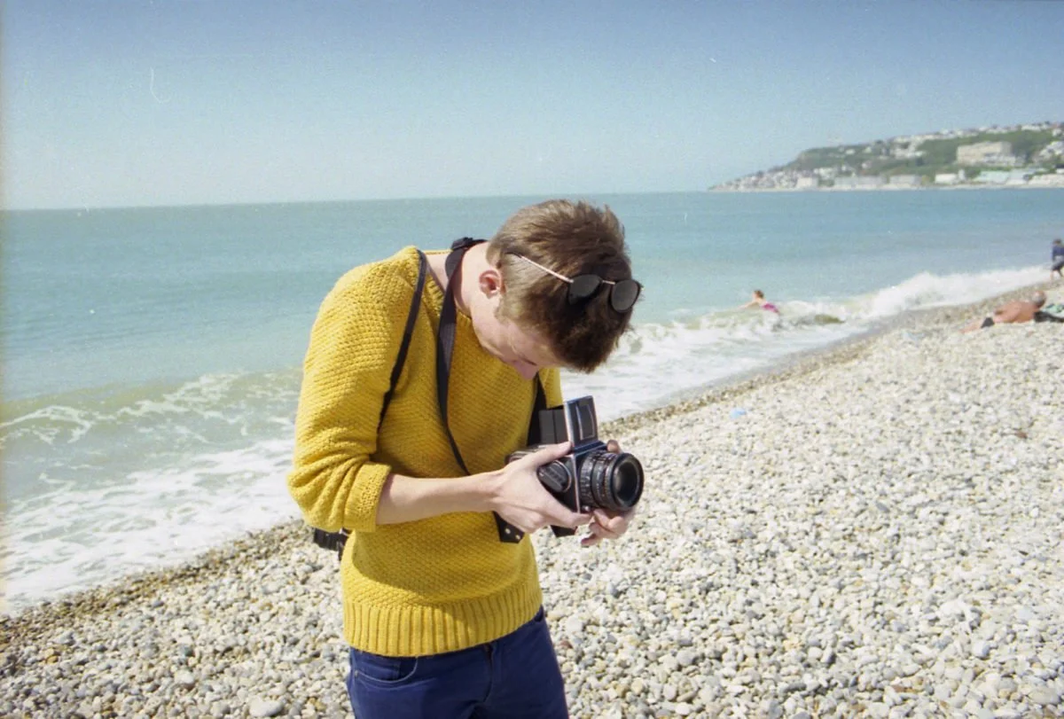 Jeune homme en pull jaune, regardant un appareil photo sur une plage de galets, avec la mer en arrière-plan.