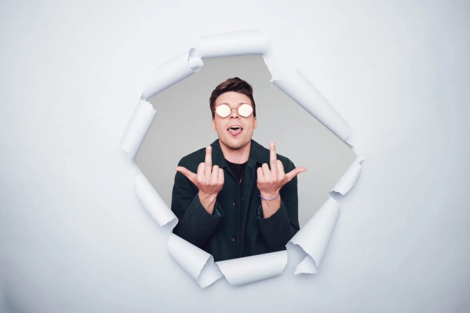 Un jeune homme avec des lunettes rondes et un blouson noir, faisant un geste obscène avec les deux mains, vu à travers un trou dans un mur blanc déchiré.