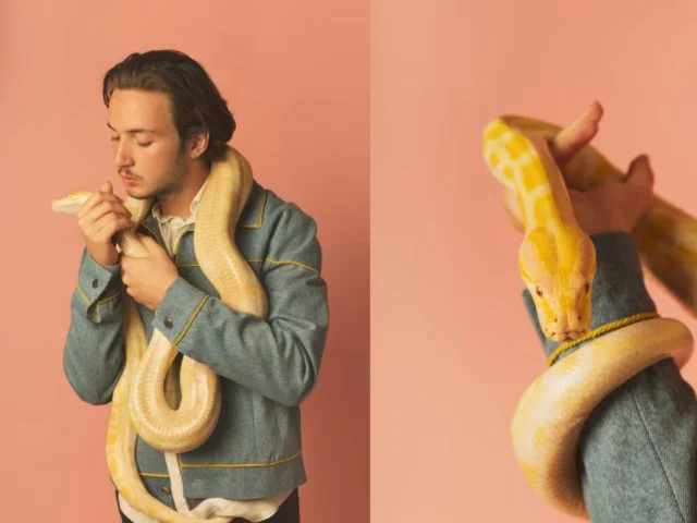 Un homme avec un serpent python jaune enroulé autour de son cou et de ses épaules, tenant une baguette de pain contre ses lèvres, sur un fond rose