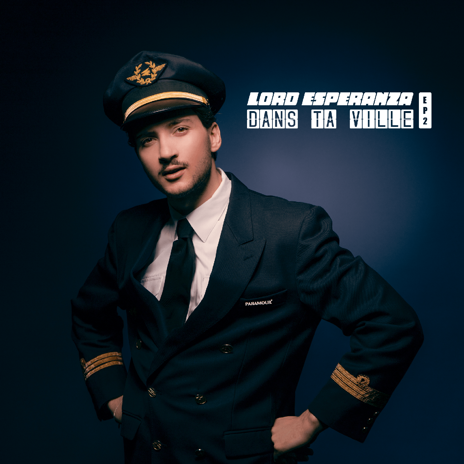 Homme en uniforme de pilote avec chapeau, veste noire avec patch 'PARAMOUR' et manches dorées, posé contre un fond sombre.