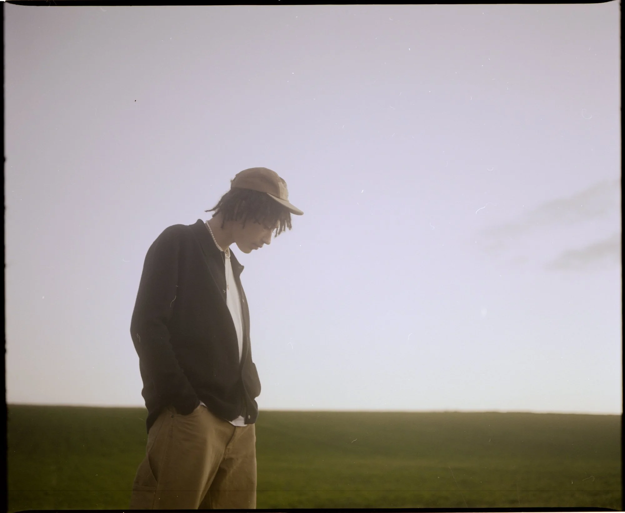Un jeune homme avec un chapeau beige et une veste noire, regardant vers le bas, debout dans un champ vert avec un ciel clair et quelques étoiles visibles.