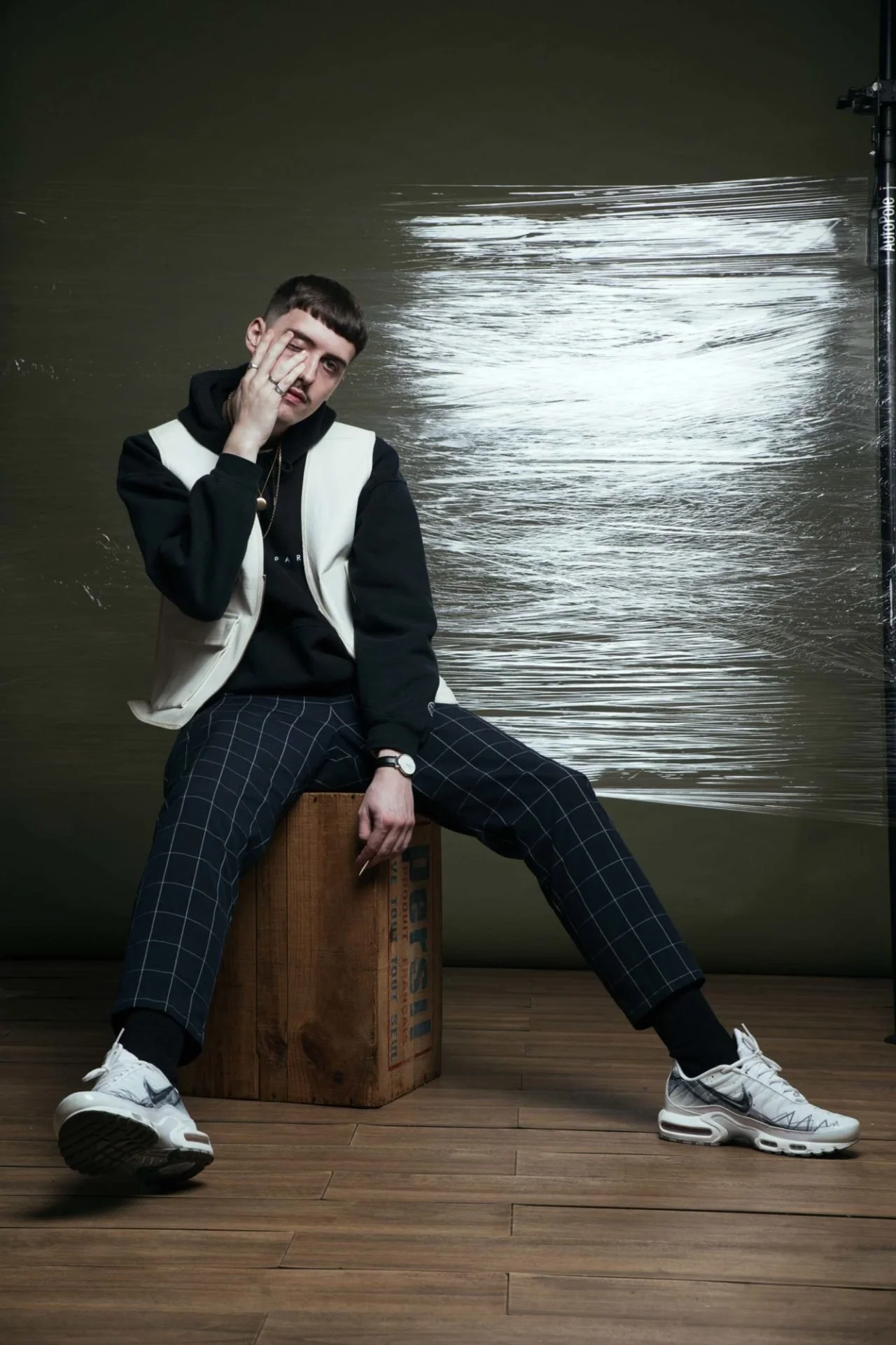Un jeune homme assis sur une caisse en bois, pose de manière décontractée devant un fond avec un effet d'eau ou de reflet. Il porte un sweat à capuche noir, un gilet blanc et un pantalon à carreaux. Il porte aussi des baskets Nike blanches et semble 
