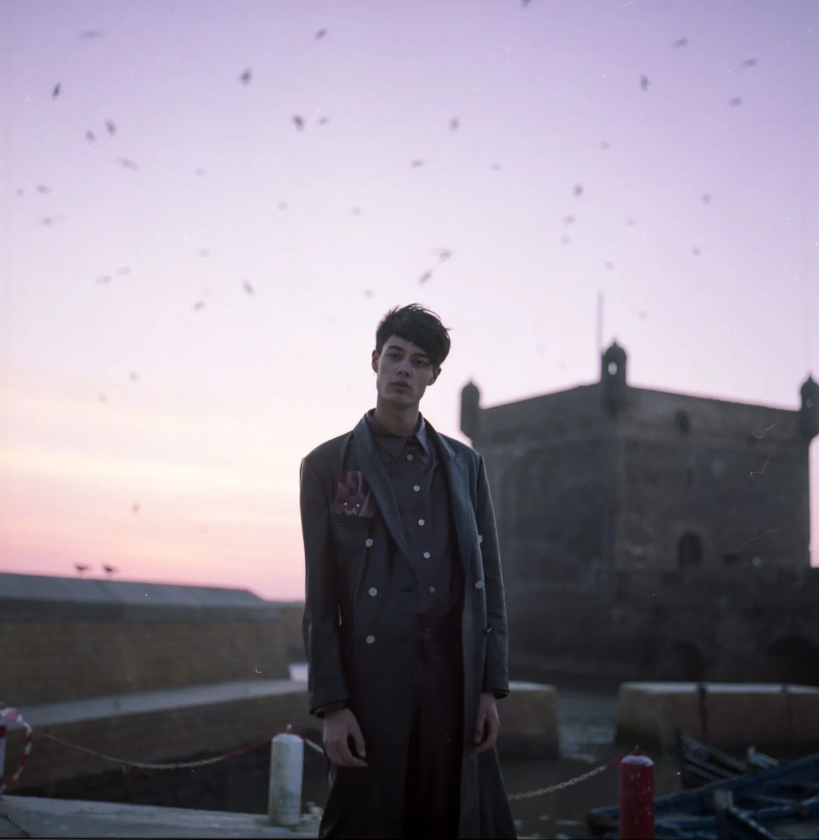 Un jeune homme debout devant un bâtiment ancien, avec un ciel crépusculaire, entouré d'oiseaux en vol.