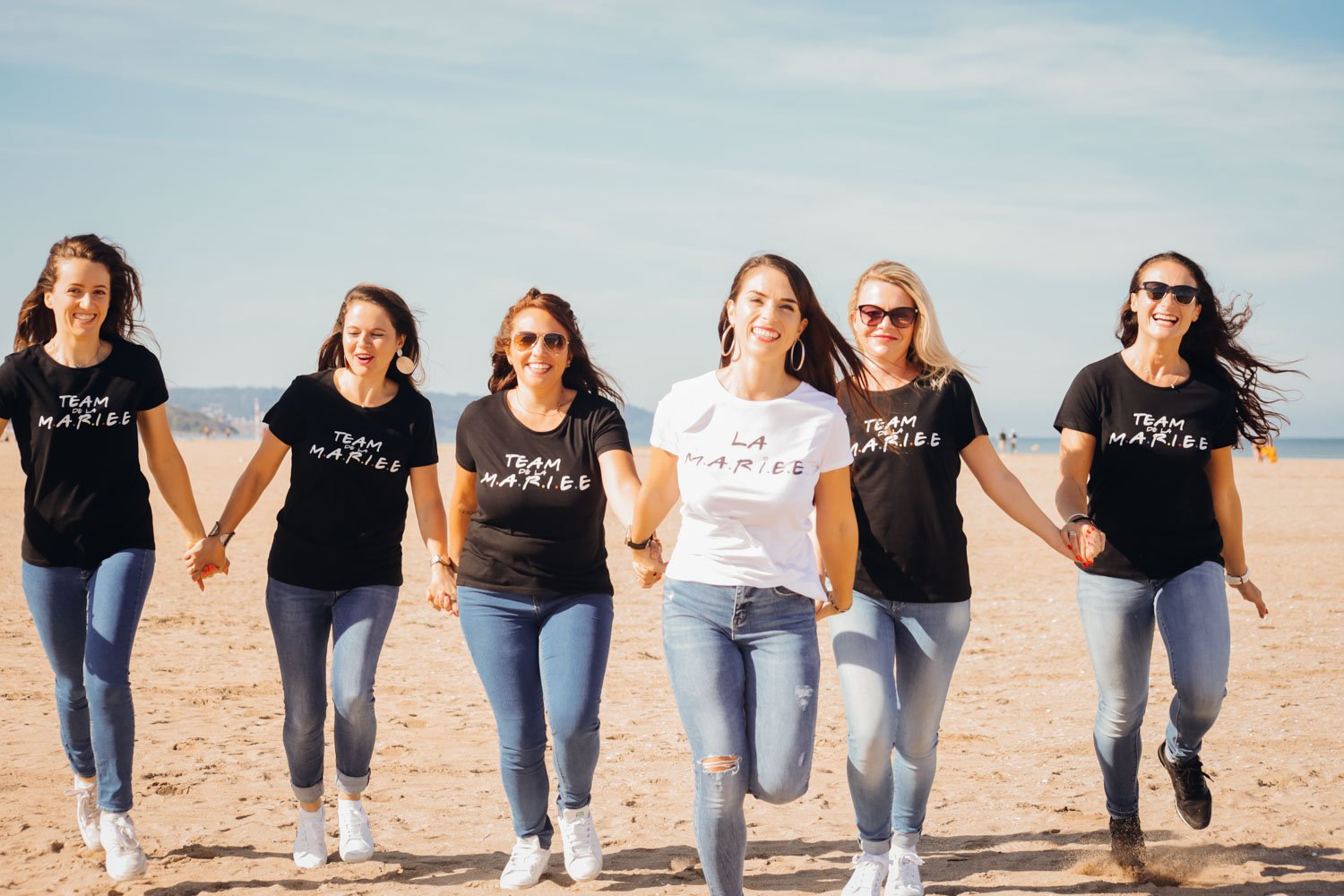 Groupe de six femmes qui s'amusent sur la plage, porteuses de t-shirts noirs ou blancs avec l'inscription "TEAM M.A.R.I.E.E" et rient tout en marchant.