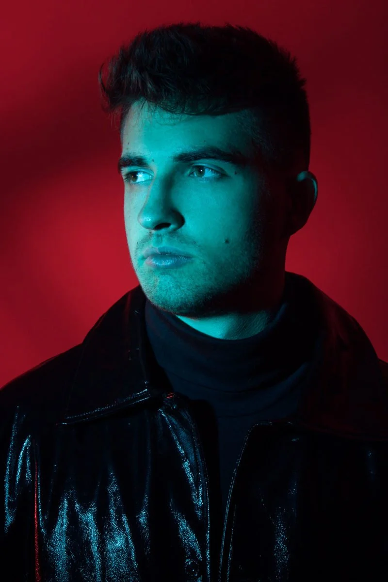 Pascal Letoublon, portant un haut noir en latex, éclairé par une lumière bleue sur un fond rouge.