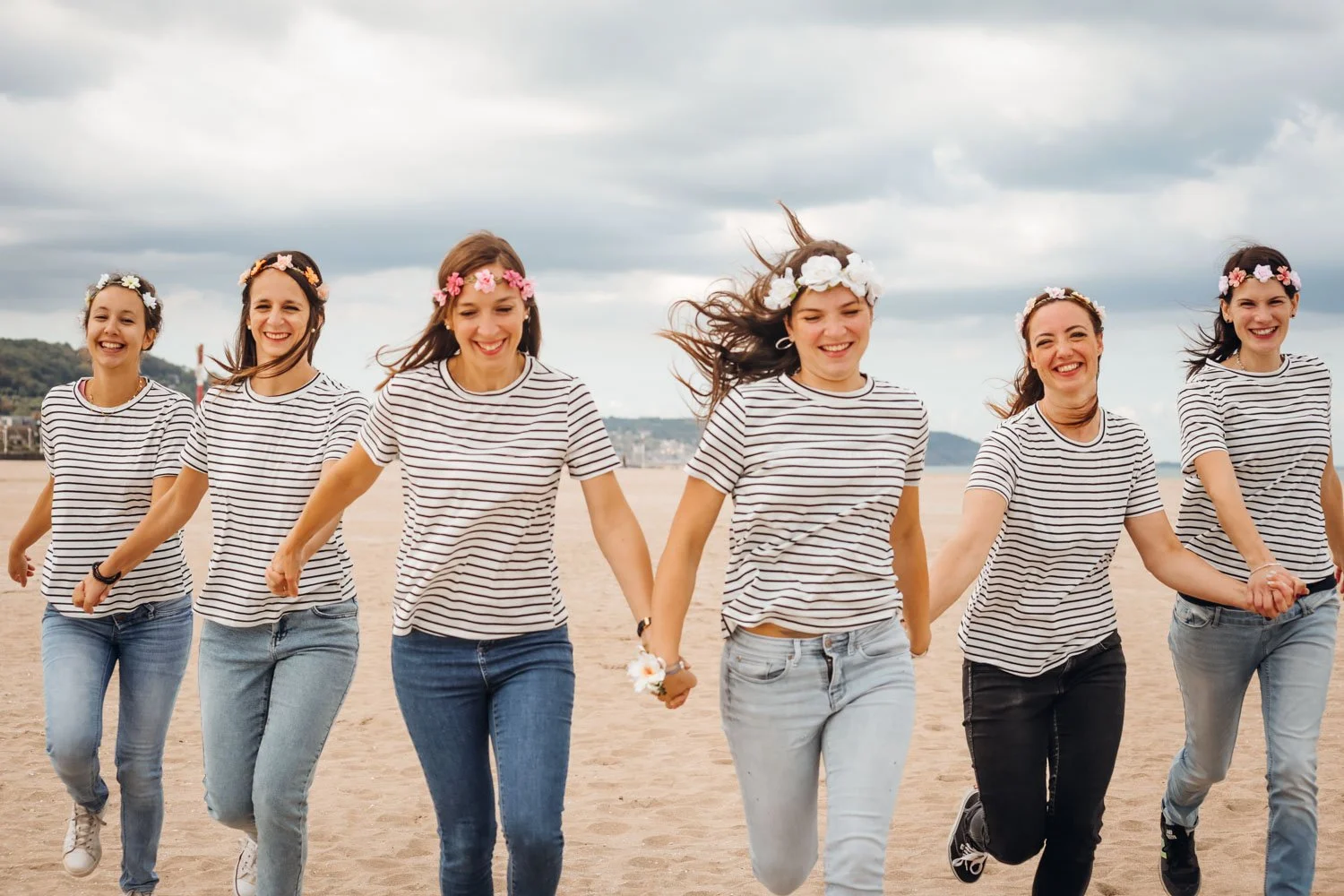 Un groupe de sept jeunes femmes souriantes, habillées en marinières, marchant joyeusement sur la plage, portant des couronnes de fleurs.