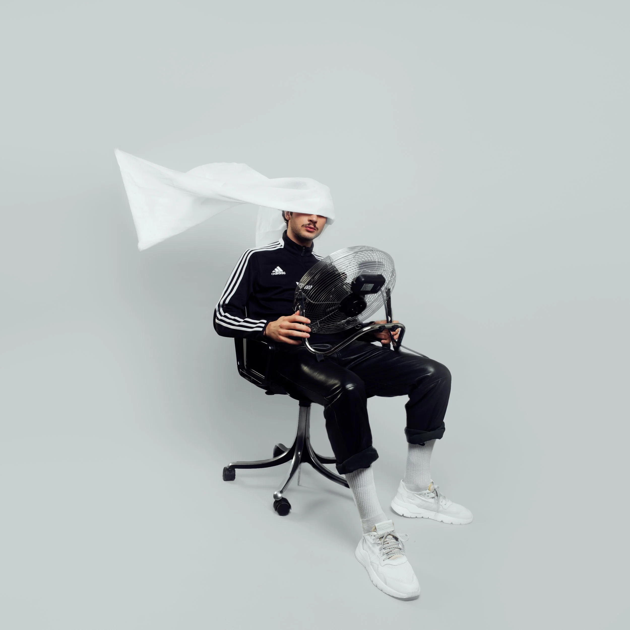 Un homme assis sur une chaise, portant un sweat à capuche noir avec des rayures blanches sur les manches, une paire de baskets blanches, une paire de chaussettes blanches, portant une paire de lunettes de soleil et tenant un ventilateur électrique. Il y a un ventilateur industriel à côté de lui et un tissu blanc flottant dans l'air, créant une sensation de mouvement ou de vent.