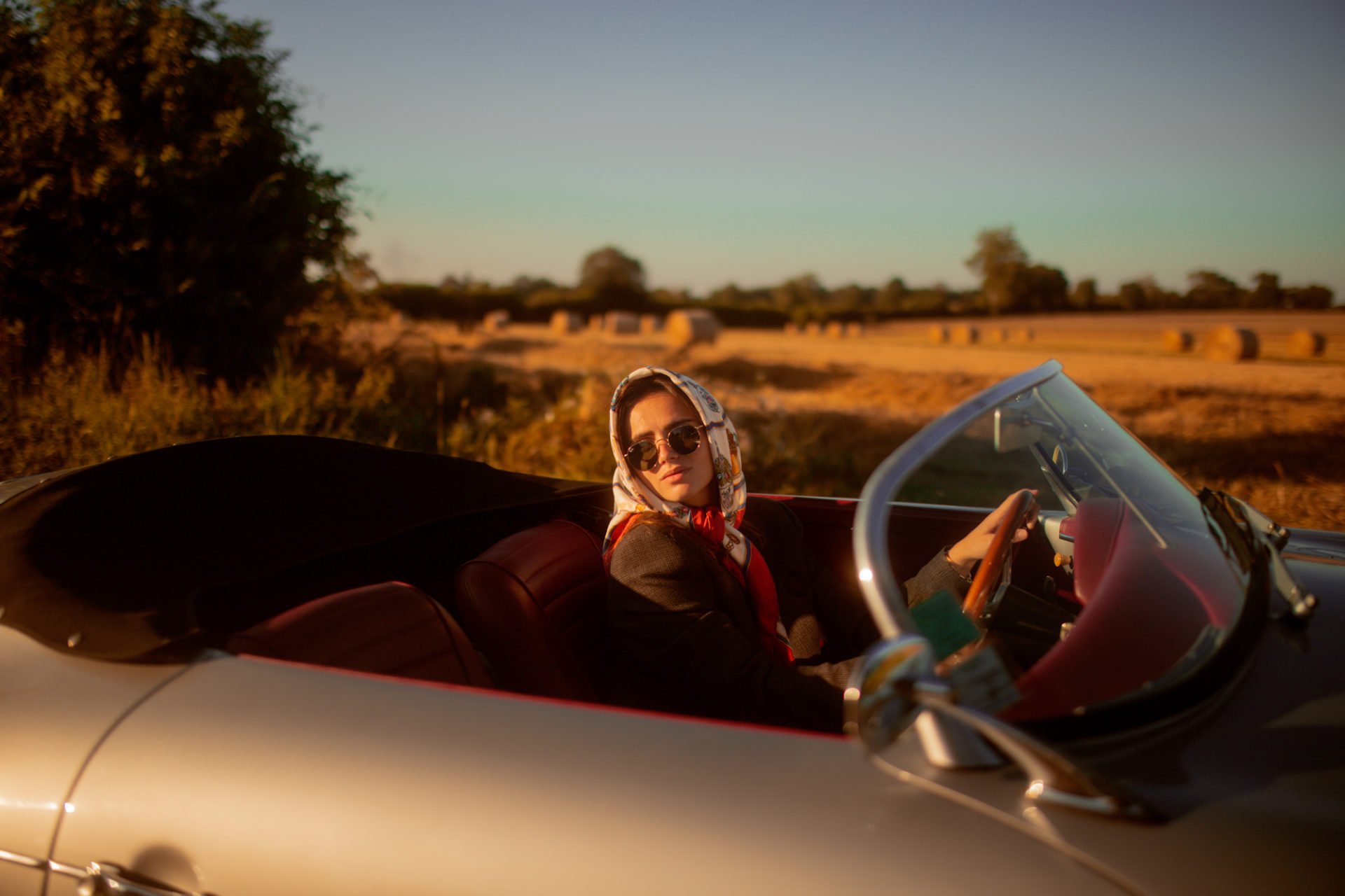 Une femme portant des lunettes de soleil et un foulard rouge dans une voiture décapotable argentée, en campagne avec des champs de foin au coucher du soleil.