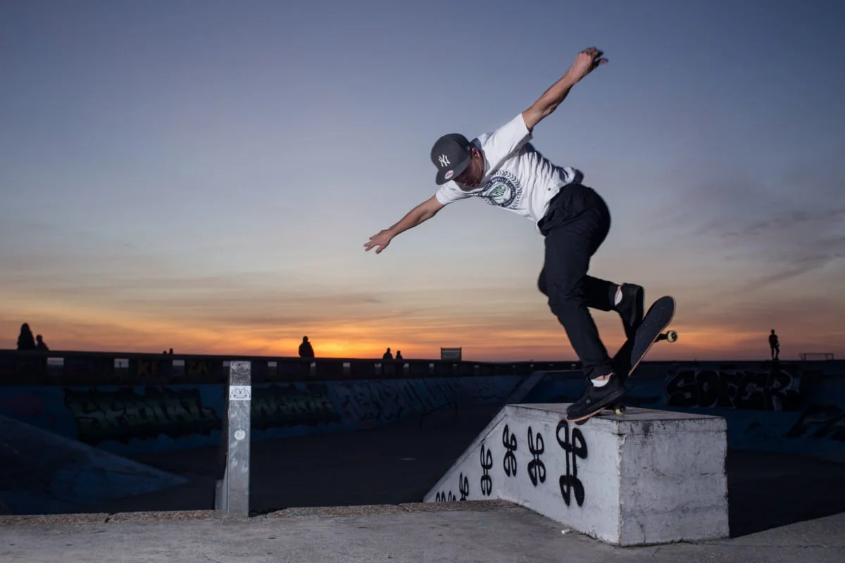 Un skateur effectue une figure sur un skate dans un skatepark au coucher du soleil, avec un ciel coloré et des silhouettes de personnes en arrière-plan.