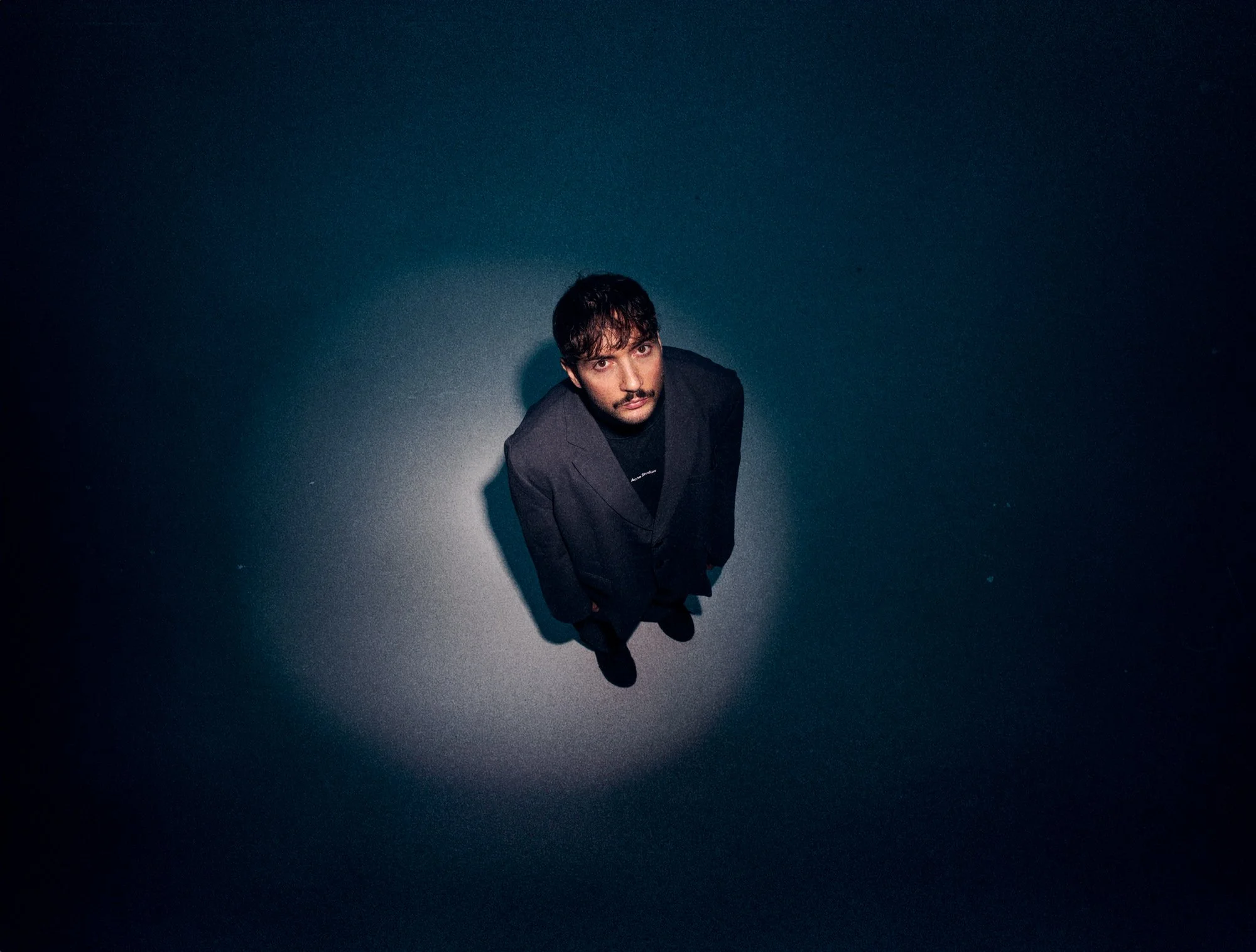 Un homme avec des cheveux bouclés et une barbe, portant un costume noir, vu de haut, se tenant devant un mur bleu sombre.