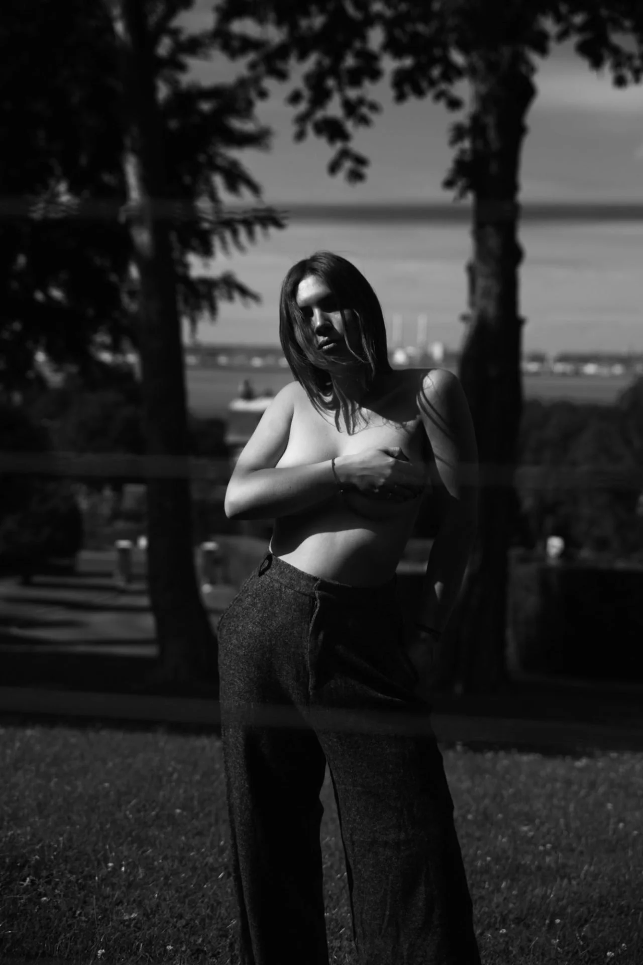 Une femme nue, portant un pantalon ample, se tient devant une clôture en fer, en pleine lumière contrastée. Arbres et bâtiments en arrière-plan, ciel clair.