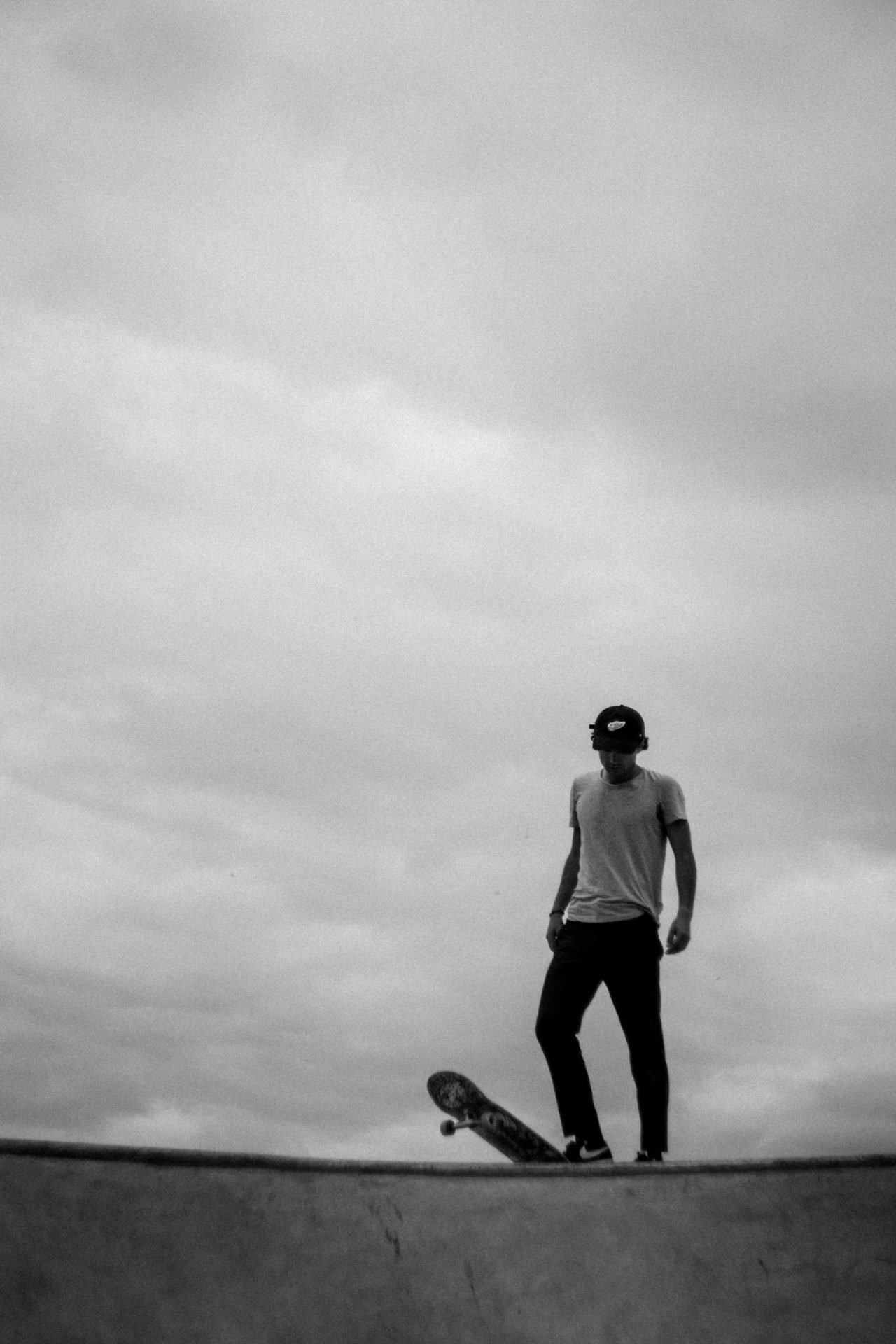 Une personne en tenue décontractée portant une casquette, qui est en train de faire du skateboard sur une surface lisse dehors, avec un ciel nuageux en arrière-plan.