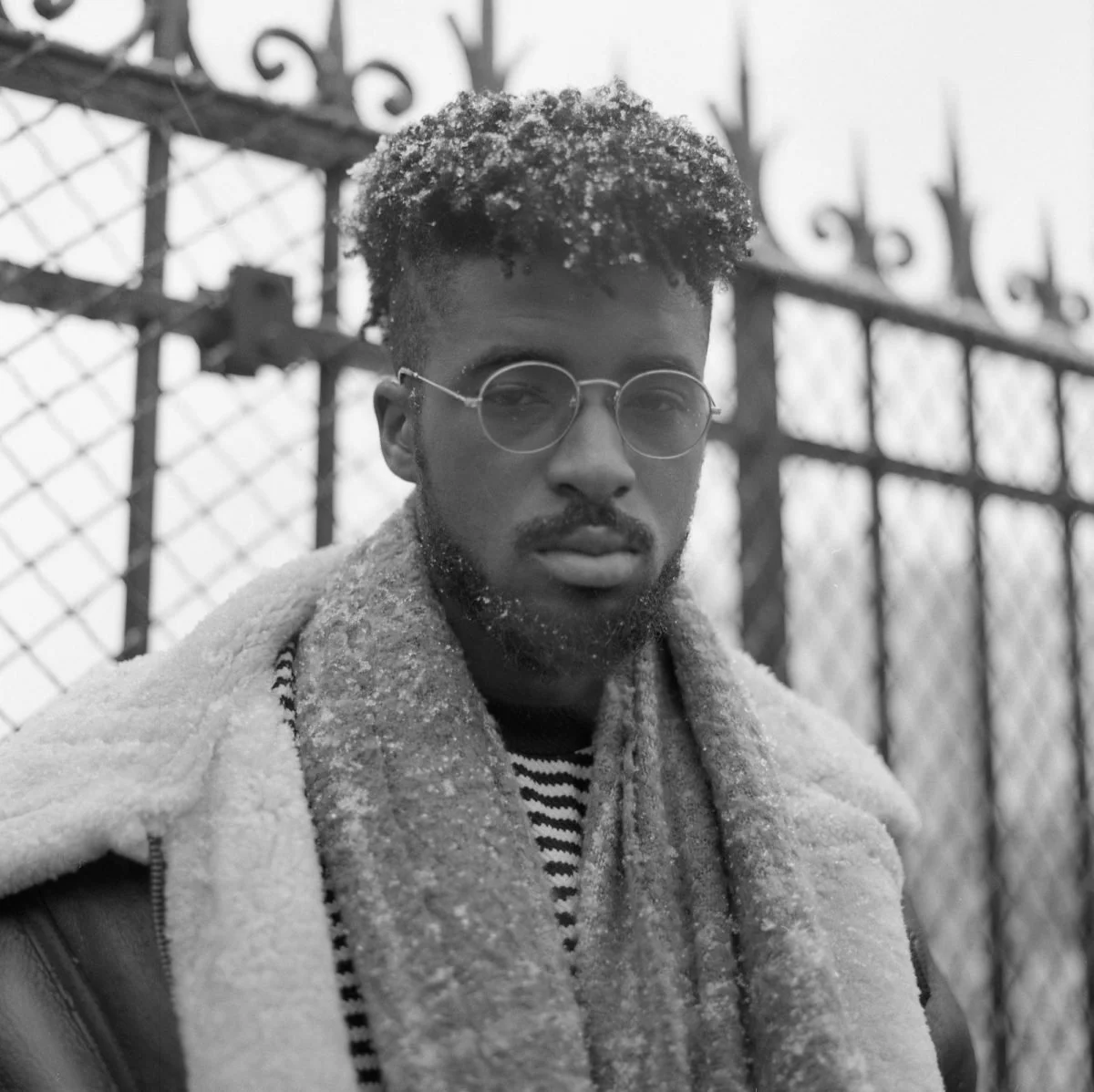 Un jeune homme avec des cheveux frisés, portant des lunettes rondes, un manteau épais et une écharpe, debout devant une grille en fer forgé par temps neigeux.