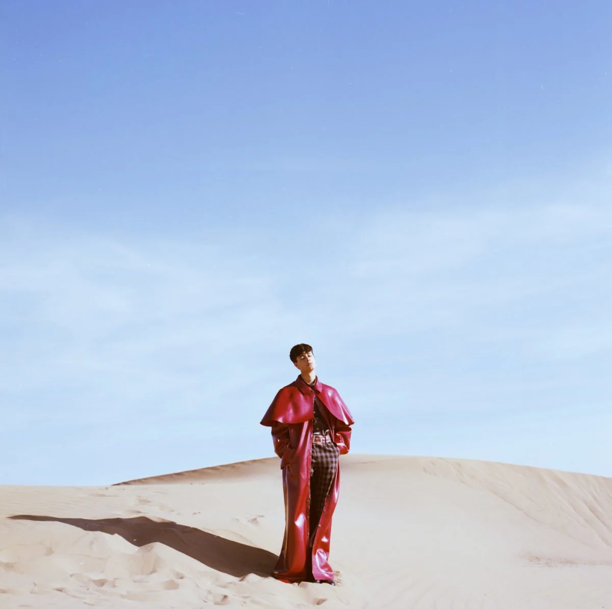 Une personne debout dans un désert de sable sous un ciel bleu clair, portant un manteau rouge et un pantalon à carreaux. Arthur Avellano
