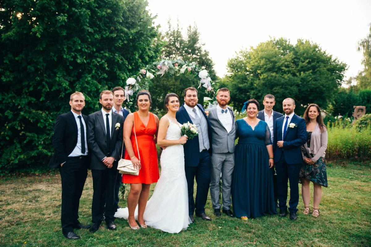 Groupe de personnes lors d'un mariage dans un jardin, la plupart portent des tenues formelles, avec la mariée en robe blanche et le marié en costume, sous une arche décorée de fleurs.