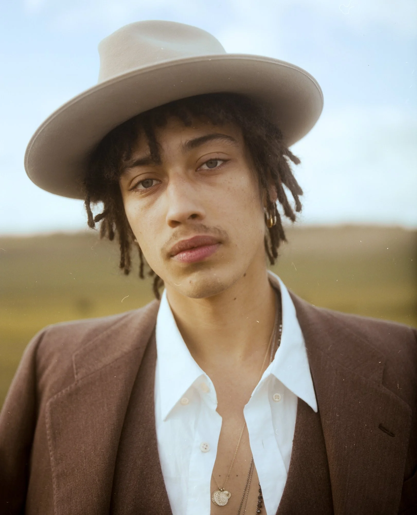Un homme portant un chapeau beige, une veste brune, une chemise blanche, avec des boucles d'oreilles et des colliers en or, dans un champ à l'extérieur.