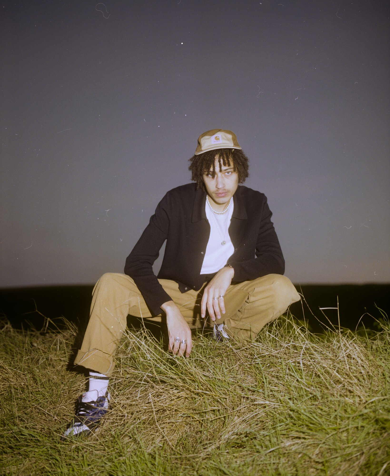 Jeune homme assis dans un champ, portant un bonnet beige, un sweat noir, un T-shirt blanc, et des pantalons beige, à la nuit avec un ciel étoilé en arrière-plan.