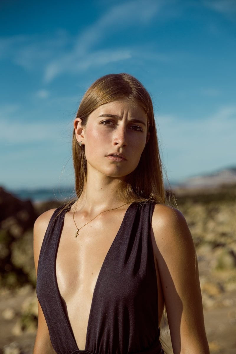Jeune femme avec cheveux longs bruns portant une robe noire à décolleté profond, debout en extérieur sous un ciel bleu avec des nuages et un paysage rocheux en arrière-plan.