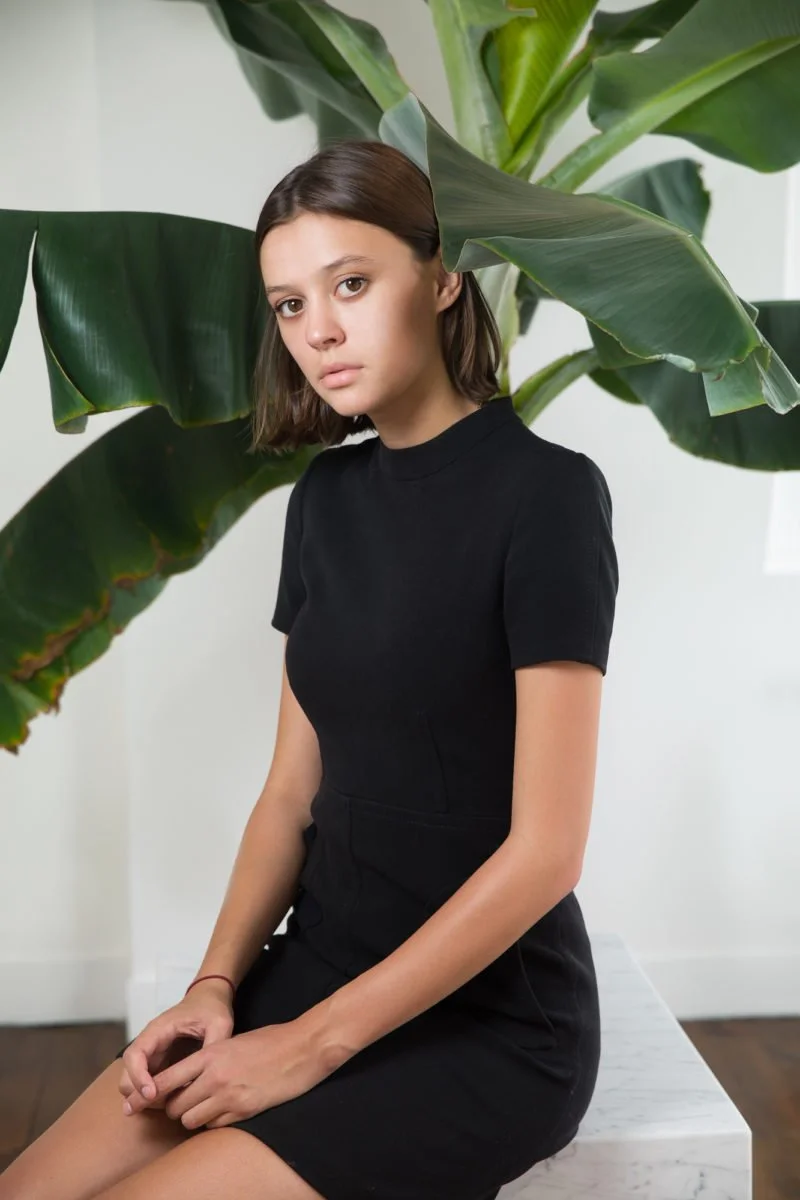 Jeune femme aux cheveux bruns courts, vêtue d'une robe noire, assise à côté d'une grande plante verte.