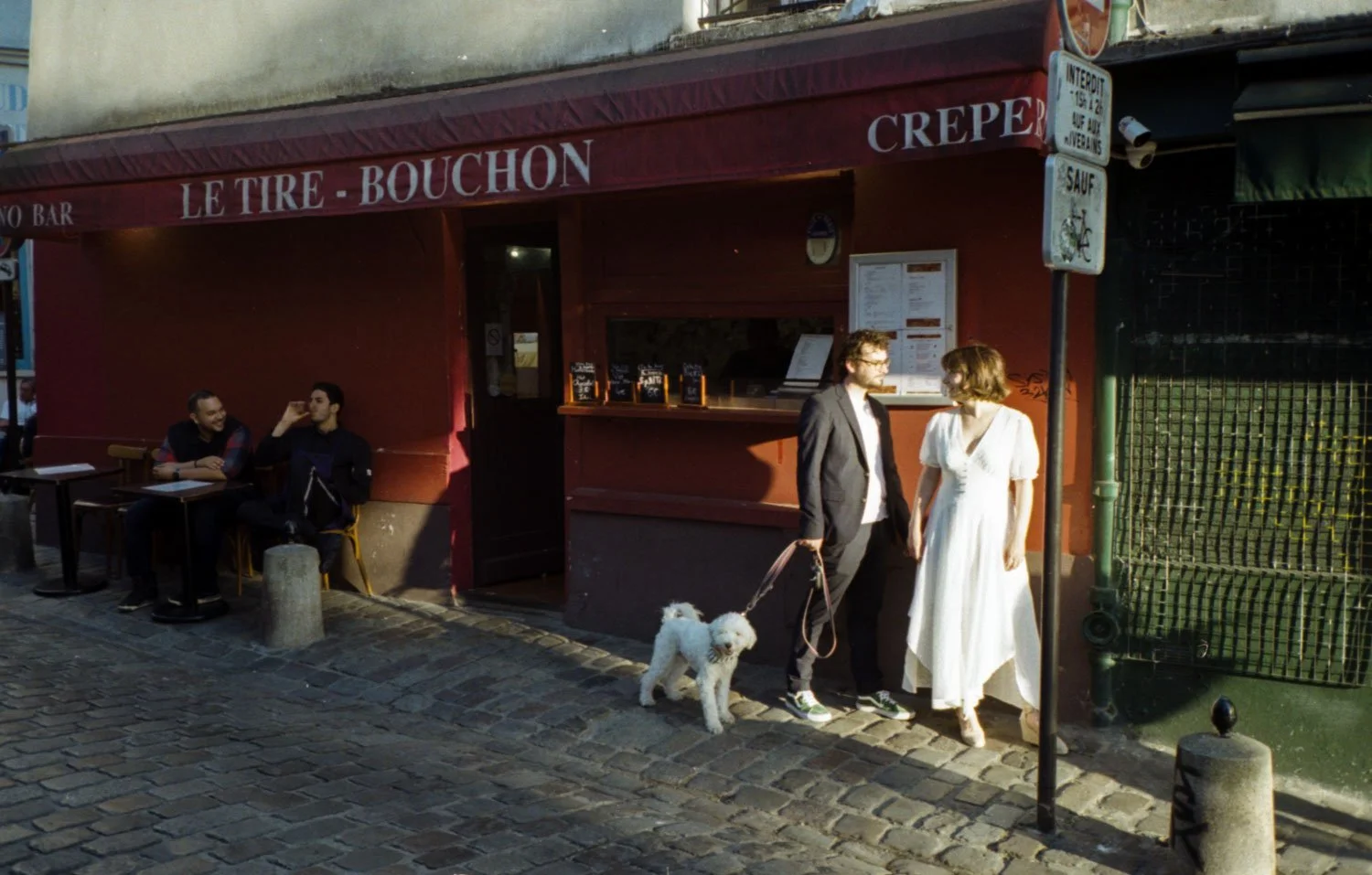 Une scène urbaine avec un restaurant nommé 'Le Tire Bouchon' à l'extérieur, où deux personnes avec un chien blanc sont en conversation, et deux autres personnes sont assises à une table en train de discuter.