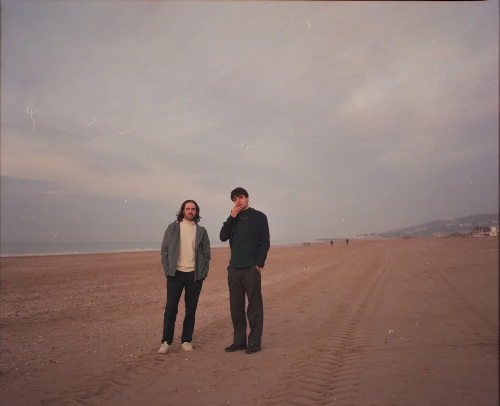 Deux hommes debout sur une plage vide, avec la mer distante et un ciel nuageux en arrière-plan.