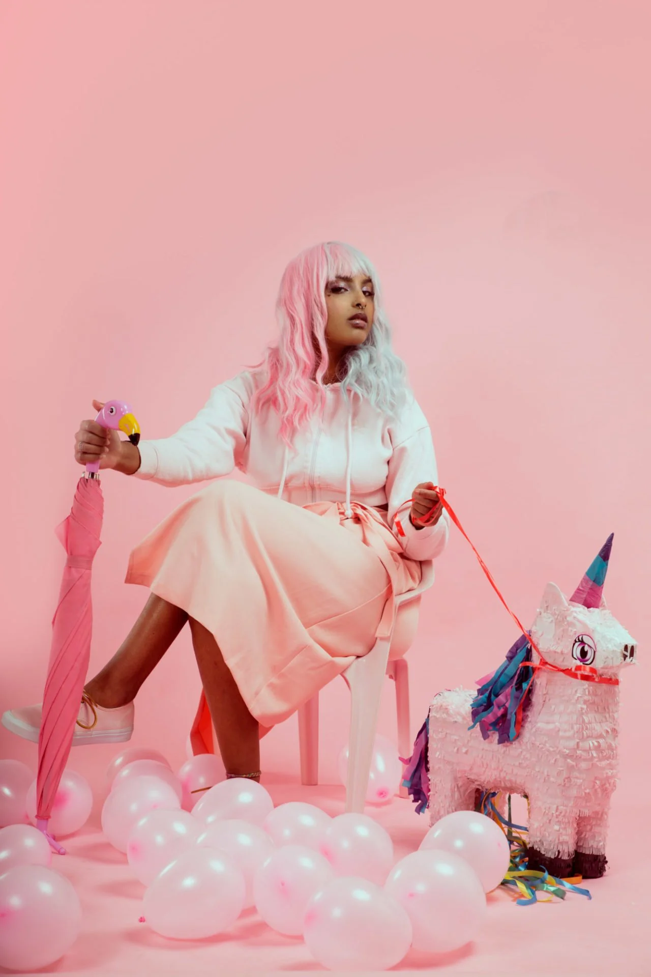 Une femme avec des cheveux mi-longs et bouclés, teint clair, portant un sweat à capuche blanc et une jupe ou pantalon rose assise sur une chaise contre un mur rose. Elle tient un flamant rose en plastique dans la main. Devant elle, il y a un piñata e