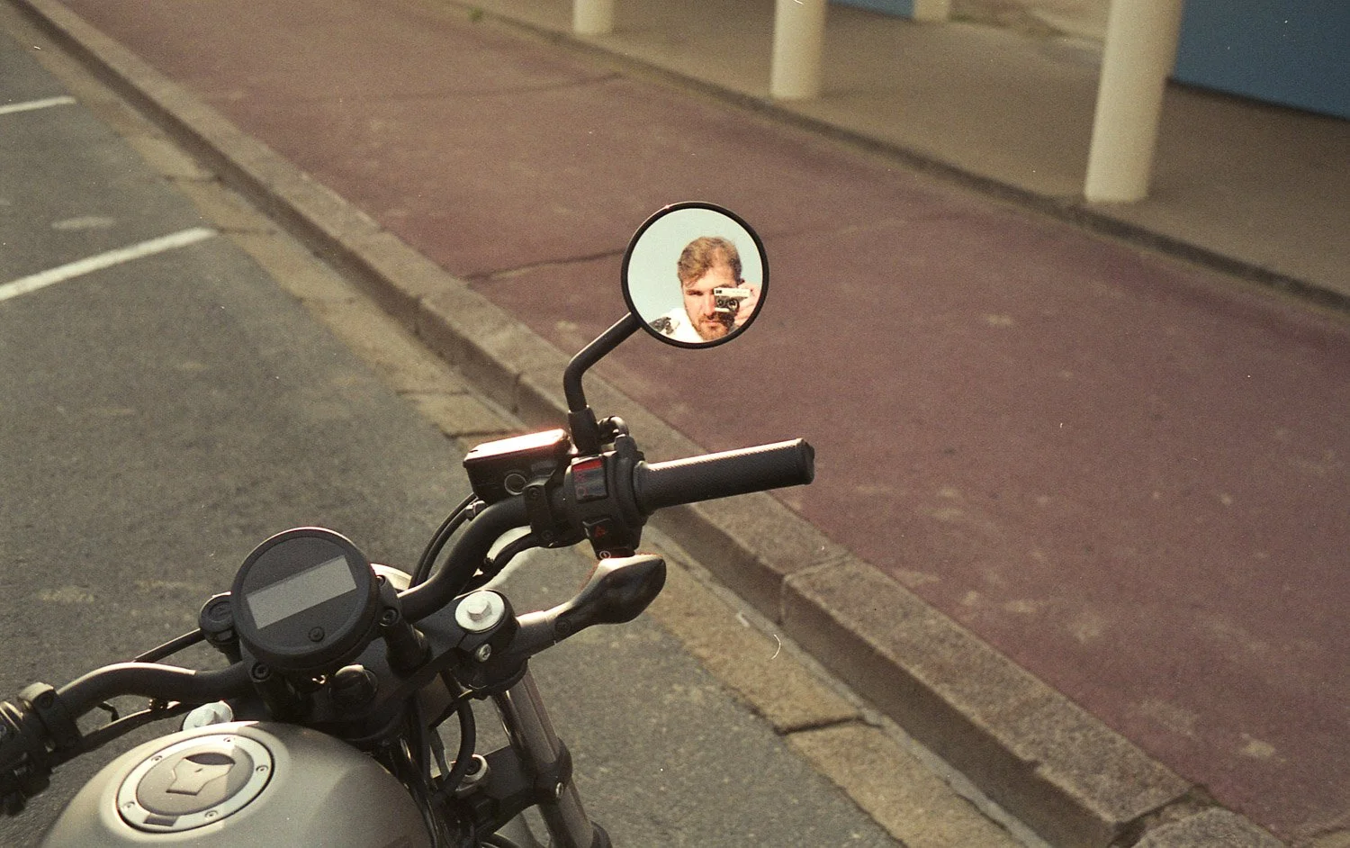 Reflection of a man with curly hair and beard taking a photo with a camera in a round mirror mounted on a motorcycle handlebar.