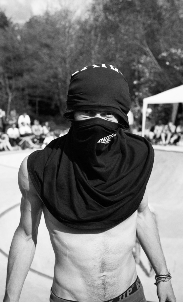 Jeune homme torse nu portant un foulard noir couvrant la majorité de son visage, avec un chapeau noir et un bandeau noir avec des écritures. Il se trouve dans un environnement extérieur avec un groupe de personnes assises en arrière-plan.