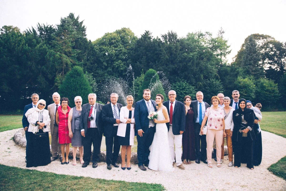 Groupe de personnes lors d'un mariage en extérieur, avec un couple de mariés au centre, entouré de famille et amis, dans un jardin verdoyant.