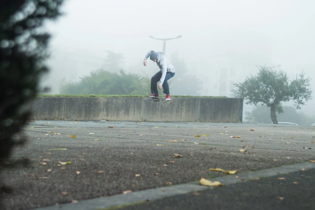 Un skateboardeur faisant un trick en dehors, sous un ciel brumeux, avec des arbres et lampadaires en arrière-plan.