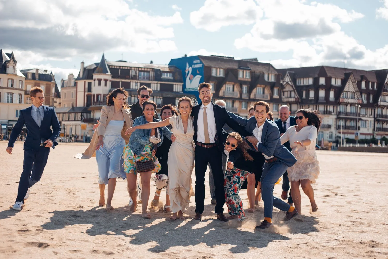 Groupe de personnes habillées formellement, joyeuses, marchant sur la plage sous un ciel en partie nuageux, avec des bâtiments en arrière-plan.