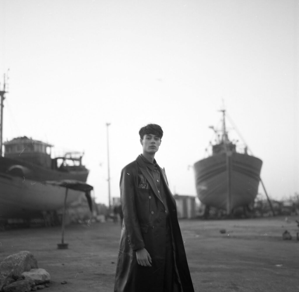 Jeune homme debout dans un port avec des navires en arrière-plan, en noir et blanc.