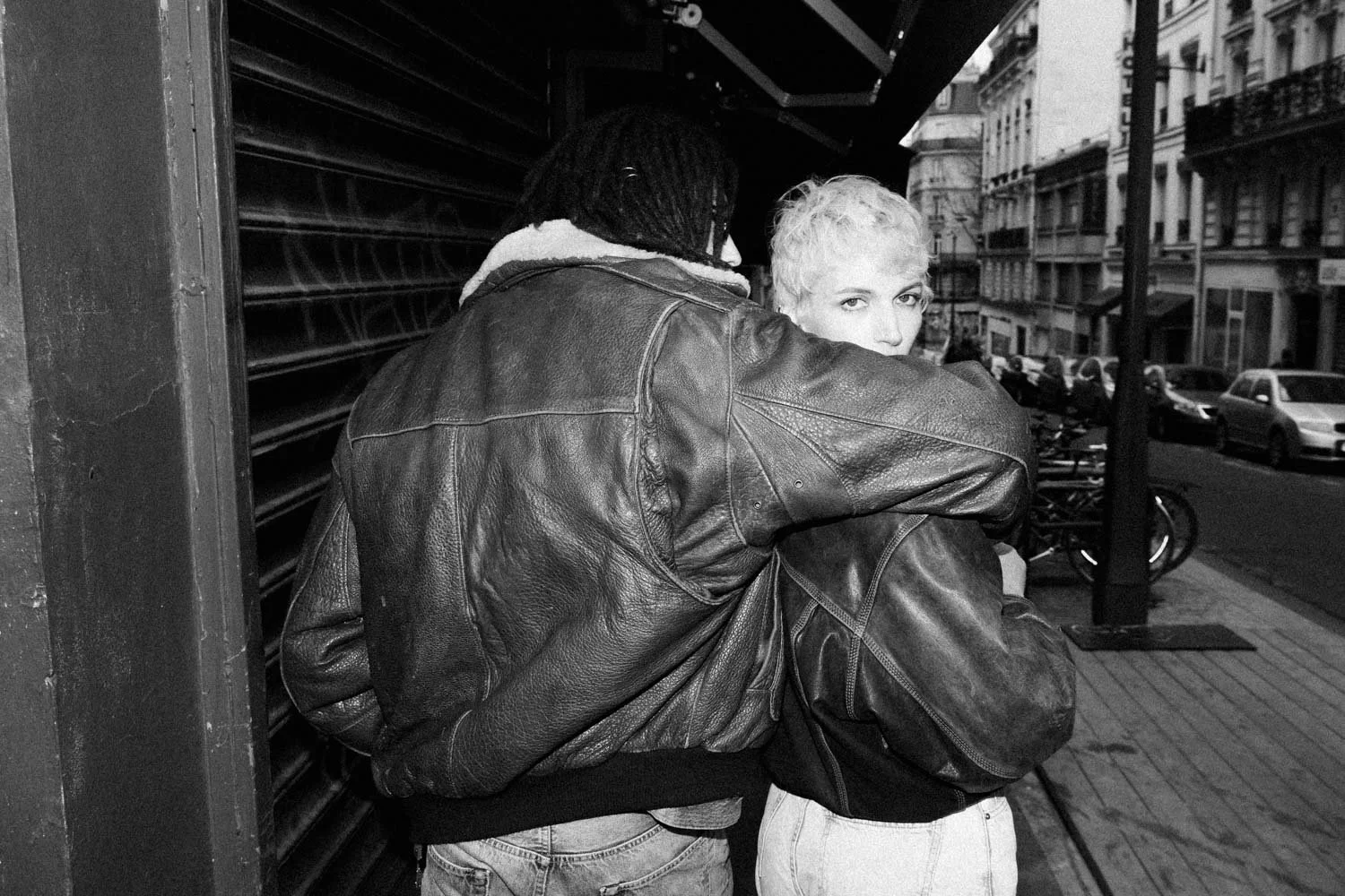 Deux personnes, dont une avec des cheveux courts et blond, se tiennent près d'un mur en ville. La personne de dos porte une veste en cuir et une écharpe, tandis que l'autre regarde vers l'objectif, sur le trottoir avec des bâtiments et des voitures e