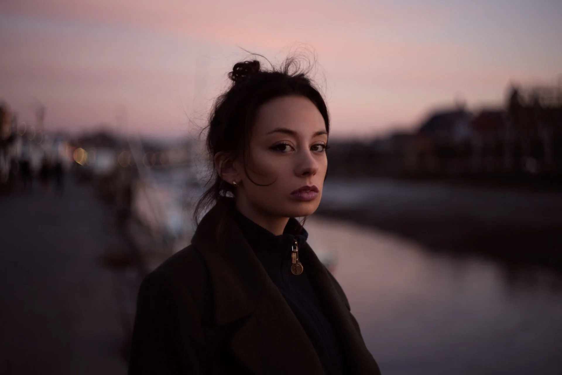 Une jeune femme aux cheveux bruns attachés, coiffée d'une petite boucle, regarde la caméra avec un fond de ciel au coucher de soleil et un paysage urbain flou.