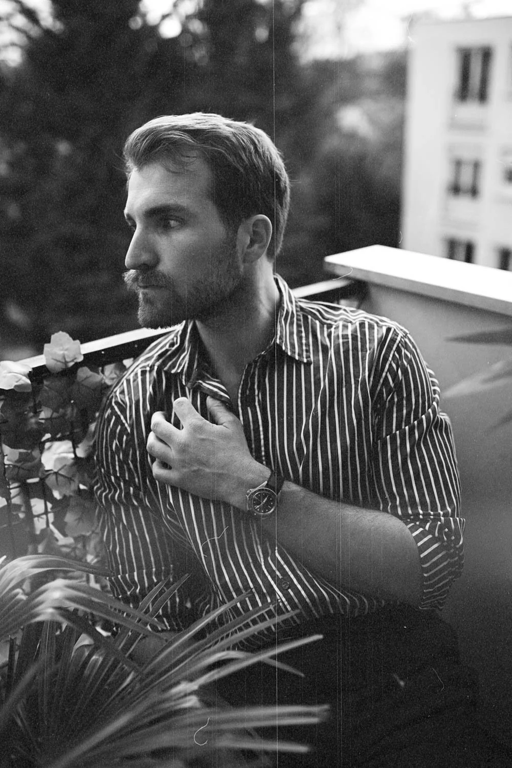 Un homme avec une barbe, portant une chemise à rayures verticales, assis sur un balcon ou une terrasse, regardant vers la gauche, avec des fleurs en premier plan