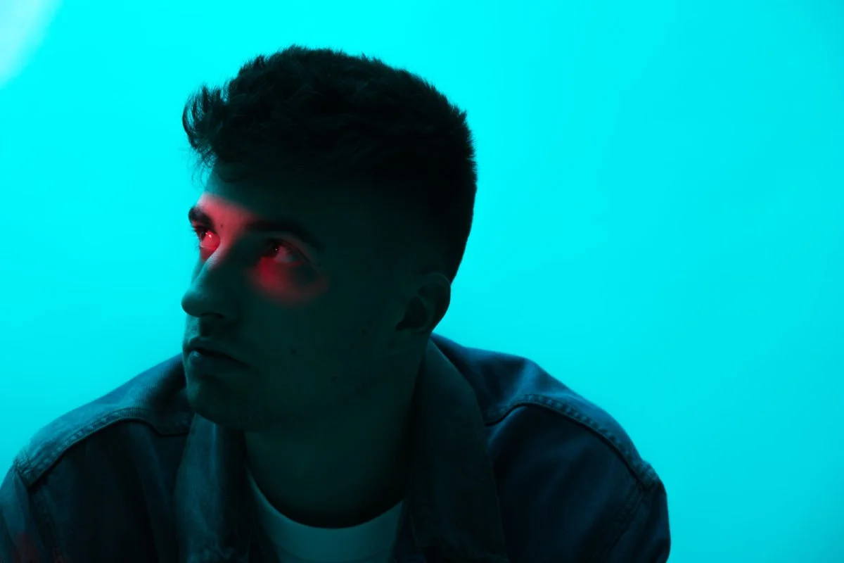 Jeune homme avec cheveux courts, regardant vers la gauche, en lumière bleue et rouge, portant une chemise en denim.