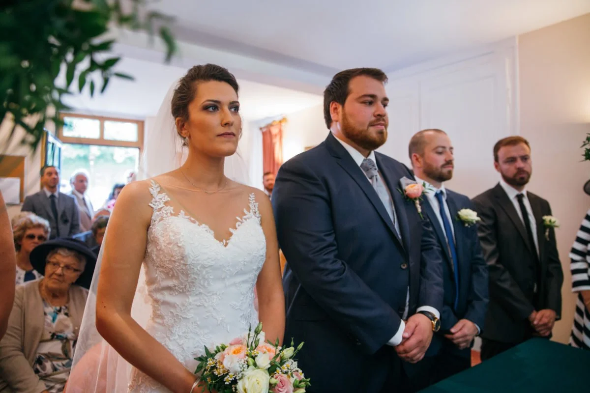 Une cérémonie de mariage avec le couple principal, la mariée en robe blanche et le marié en costume, entourés de leurs témoins et invités dans une pièce lumineuse.