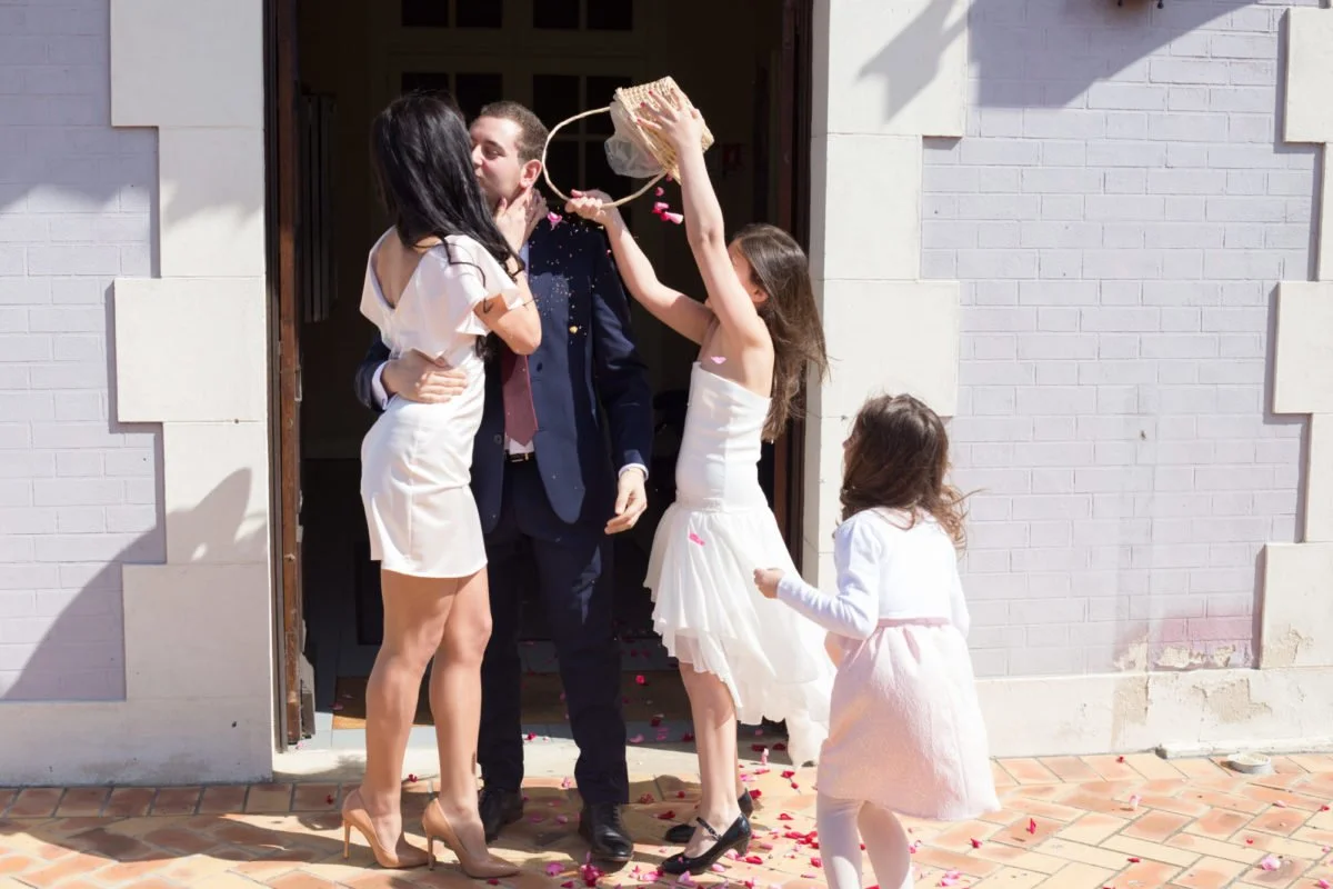 Une famille sortant d'une maison en célébration, avec des pétales de fleurs et confettis, une femme et deux jeunes filles en robe blanche, un homme en costume, un des jeunes filles verse de l'eau sur un homme, célébration joyeuse.