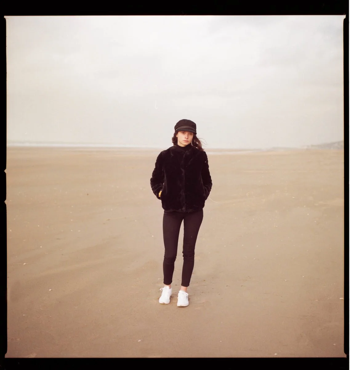 Une jeune femme en manteau noir, bonnet et baskets blanches, marche sur une plage désertique par une journée nuageuse.