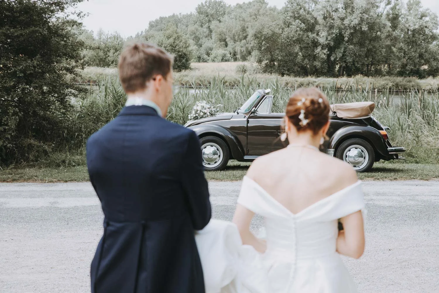 Un couple en vêtements de mariage regardant une voiture décapotable noire décorée, dans un cadre naturel avec des arbres et un étang en arrière-plan.