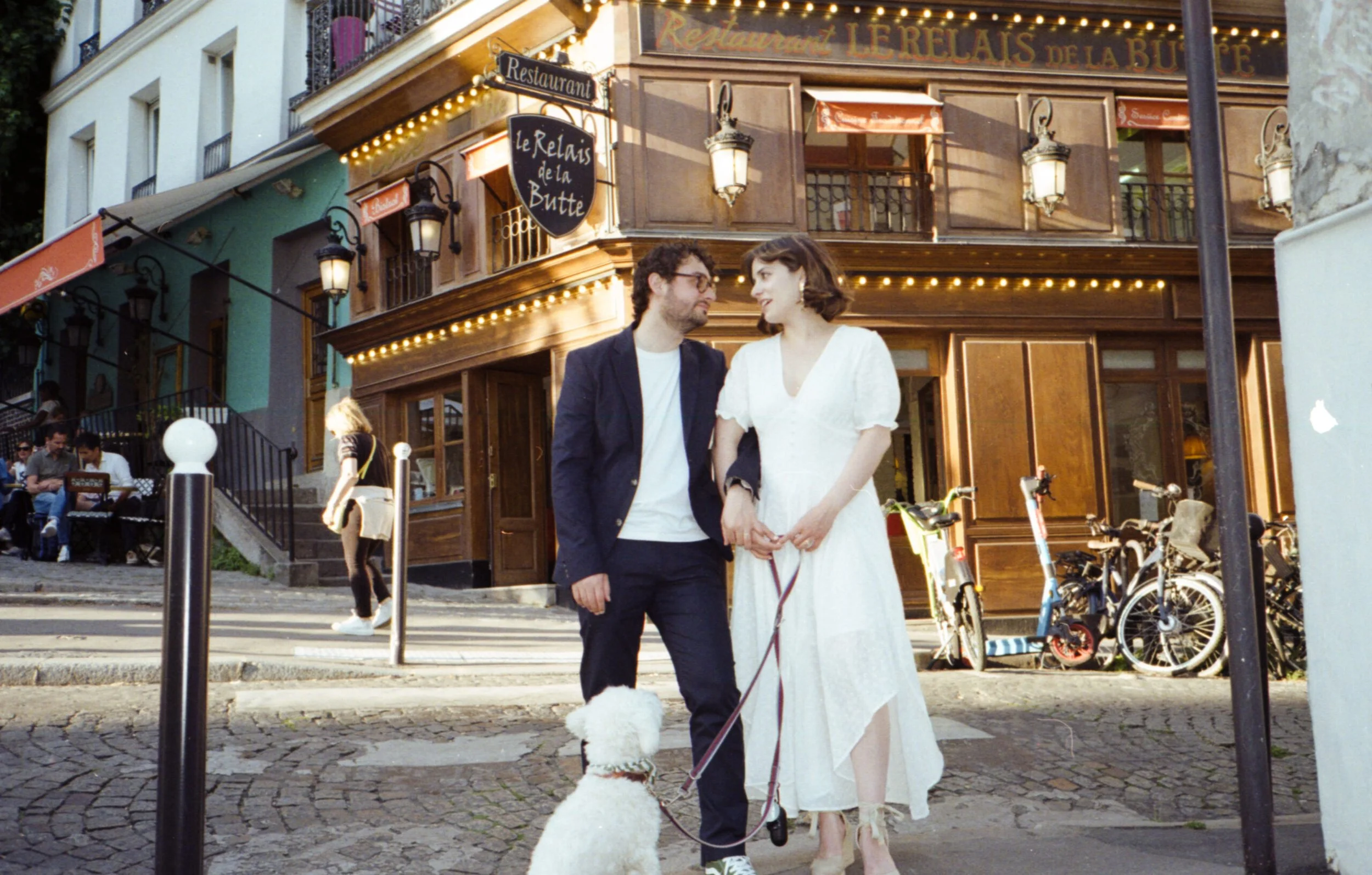 Un couple habillé élégamment,tenant la main, se tenant devant un restaurant appelé 'Le Relais de la Butte' à Paris. Le restaurant est décoré de lumières chaudes et possède une façade en bois avec des lampes anciennes. Il y a des vélos garés sur le tr