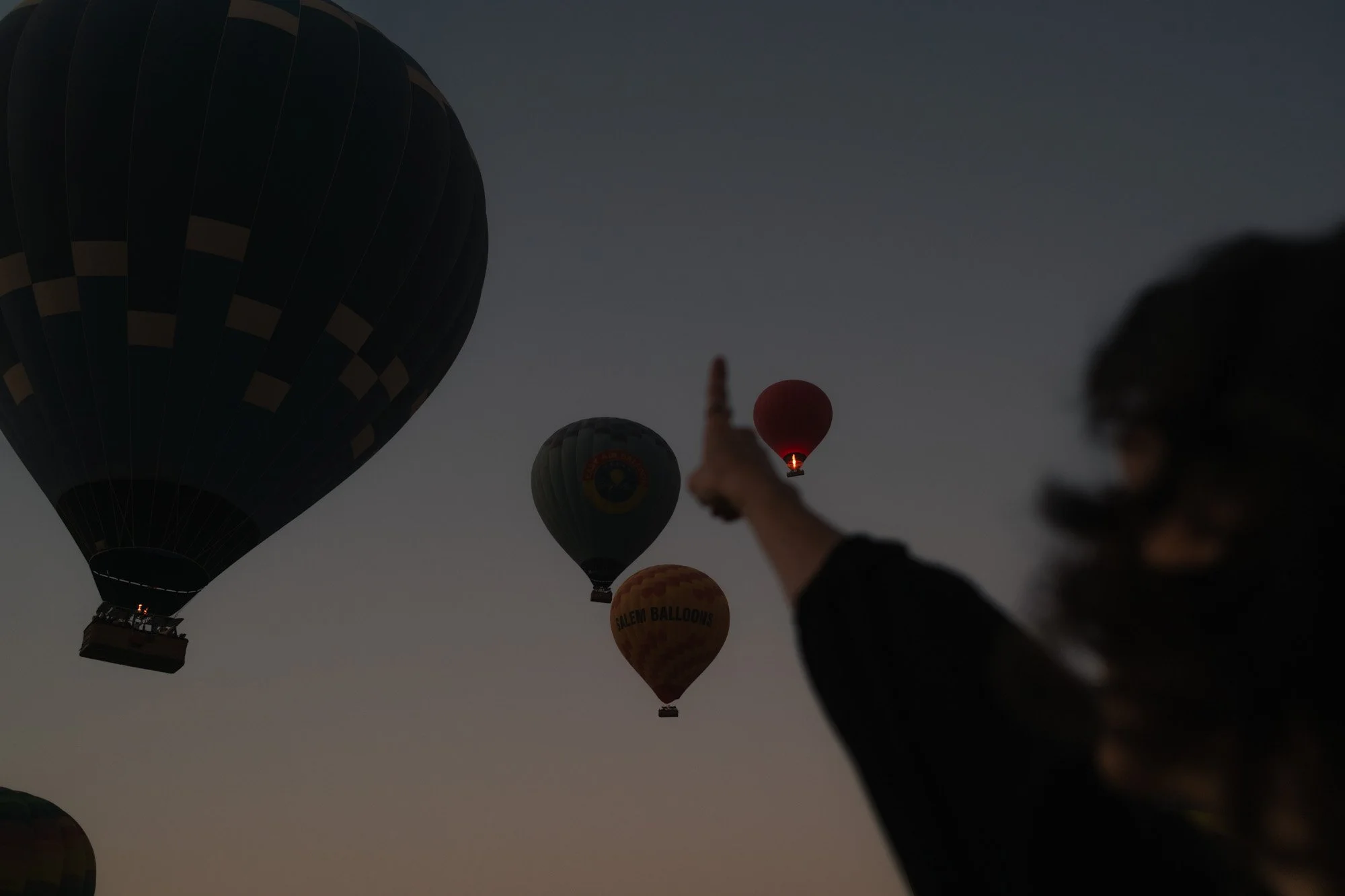 Plusieurs montgolfières colorées flottent dans le ciel au coucher du soleil, avec une personne en premier plan pointant vers elles en egypte