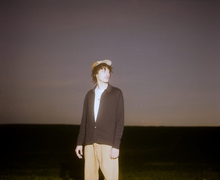 Jeune homme avec un chapeau, des vêtements décontractés, se tenant dans un paysage nocturne avec un ciel étoilé.