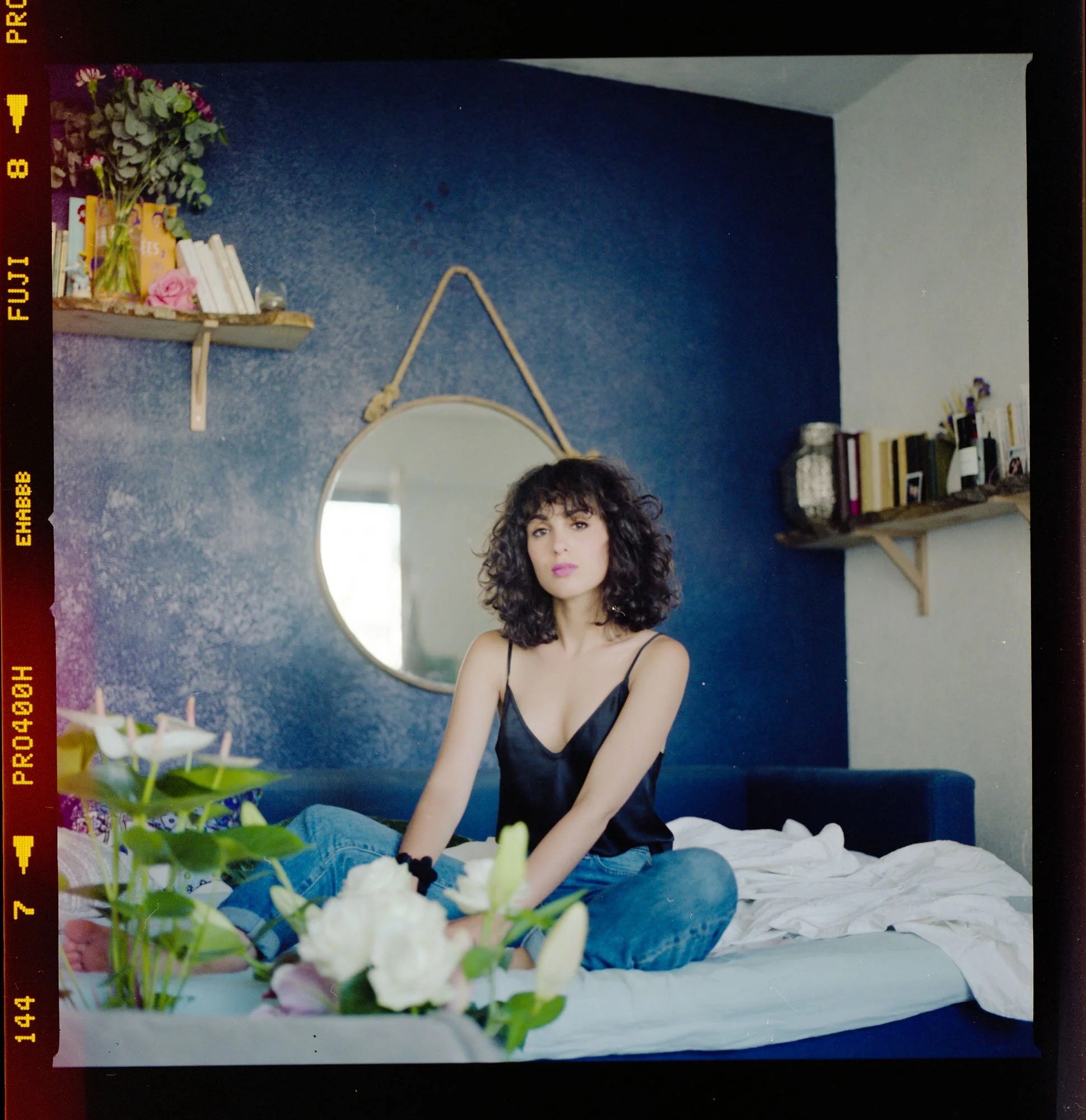 Une femme aux cheveux bouclés, portant un haut noir, assise sur un lit blanc avec des fleurs blanches devant elle. La pièce a un mur bleu avec une étagère en bois contenant des livres, des plantes et des décorations.