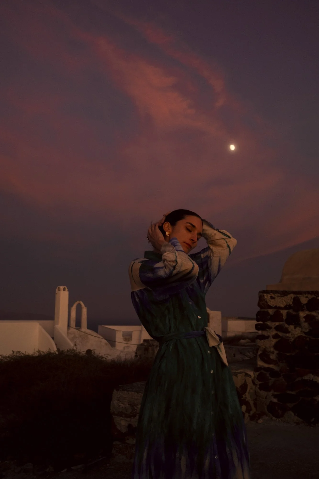 Femme debout la tête inclinée avec mains dans les cheveux, sur un toit de nuit avec ciel étoilé, lune visible, architecture blanche en arrière-plan à santorin