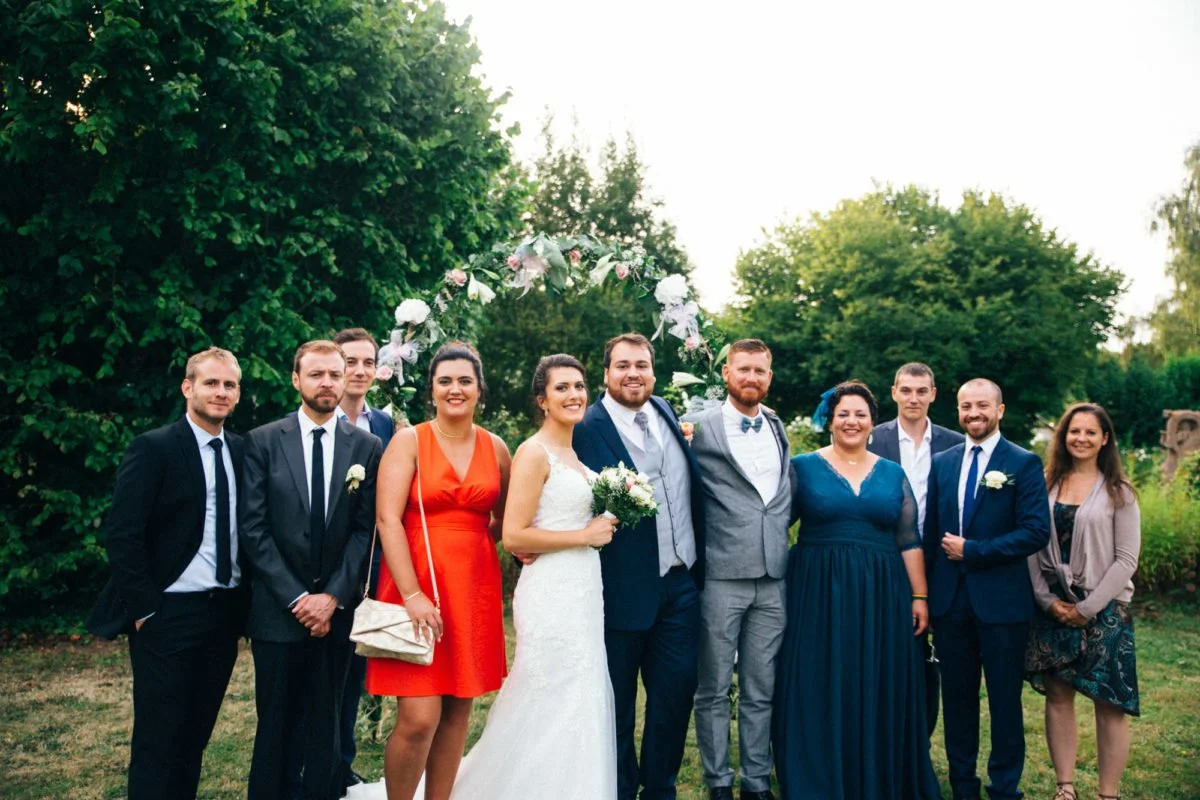 Groupe de dix personnes lors d'un mariage, avec un couple nuptial au centre, entouré d'amis et de famille, en extérieur avec des arbres en arrière-plan, sous une arche de fleurs.